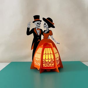 Pode incluir: Uma peça de artesanato em papel com tema de Halloween, apresentando um casal de esqueletos em trajes formais, com uma lanterna laranja decorativa em primeiro plano. A lanterna tem um design intrincado e emite um brilho quente. Os esqueletos estão em pose de dança.