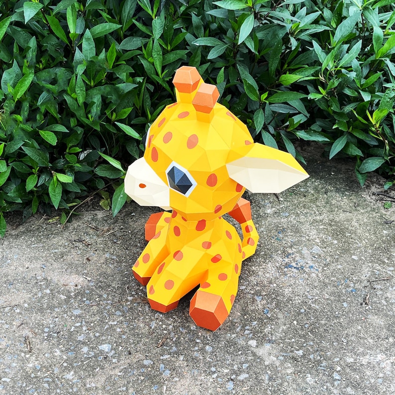 May include: A yellow and orange papercraft giraffe with black spots. The giraffe is sitting on a grey concrete surface.