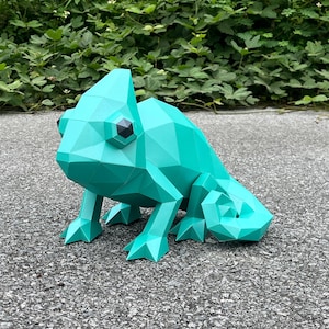Op de afbeelding: Een turquoise geometrische papieren kameleonsculptuur met zwarte ogen. De kameleon staat op een grijze ondergrond met groene bladeren op de achtergrond. De sculptuur heeft een polygonale vormgeving en is een uniek decoratief item.