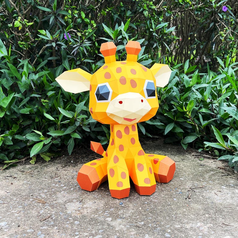 May include: A yellow and orange papercraft giraffe with brown spots, sitting on a grey surface. The giraffe has large, black eyes and a pink nose.
