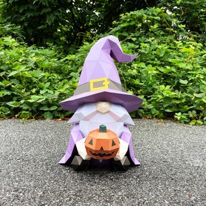 Può includere: Uno gnomo di Halloween di carta con un cappello da strega viola con fibbia dorata e mantello abbinato. Lo gnomo tiene in mano una zucca arancione geometrica con un gambo verde. Lo gnomo è su una superficie grigia con fogliame verde sullo sfondo.