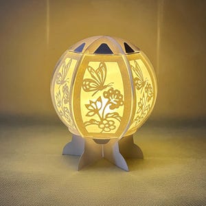 以下が含まれることがあります： 蝶と花がくり抜かれた丸いデザインの白い紙のランプ。ランプは点灯しており、温かい光を放っています。