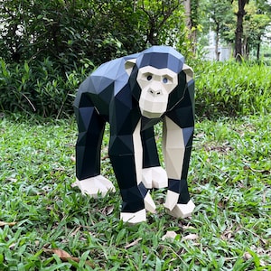 Könnte beinhalten: Eine geometrische Papierskulptur eines Schimpansen, hauptsächlich in Schwarz und Weiß, steht auf grünem Gras. Der Affe ist im Low-Poly-Stil dargestellt, mit winkligen Ebenen, die seine Form definieren. Die Skulptur befindet sich im Freien, mit Bäumen und Laub im Hintergrund.
