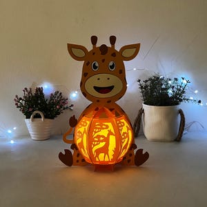 May include: A brown and orange papercraft giraffe lamp with a light inside. The giraffe is holding a round, orange, papercut lampshade with a giraffe silhouette design. The lamp is sitting on a white surface.