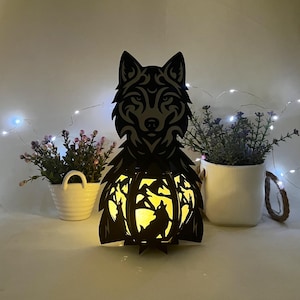 May include: A black, laser-cut wooden wolf lantern with a detailed wolf head design. The lantern features a forest scene with a wolf silhouette when lit. The lantern is surrounded by small potted plants and fairy lights, creating a warm, decorative ambiance.