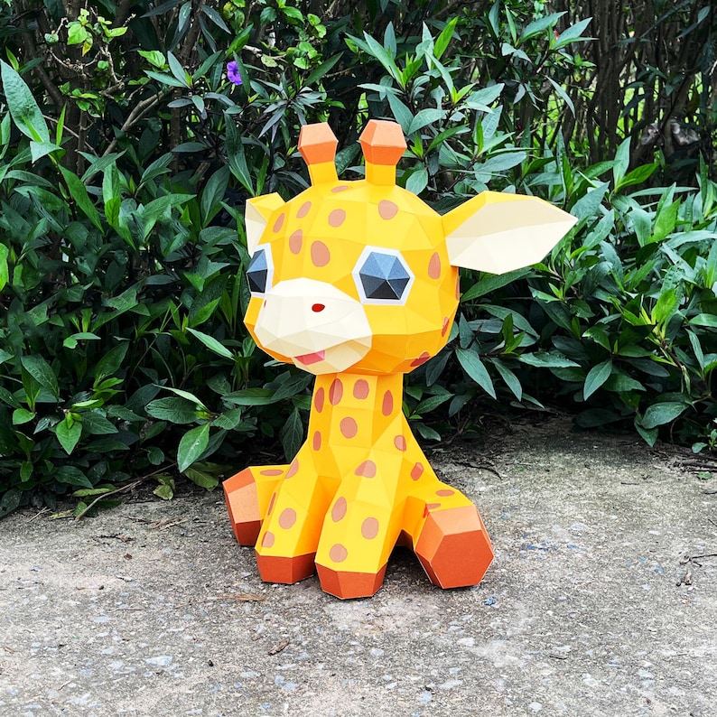 May include: A yellow and orange papercraft giraffe with black spots. The giraffe has large eyes and a friendly expression. The giraffe is sitting on a grey concrete surface.