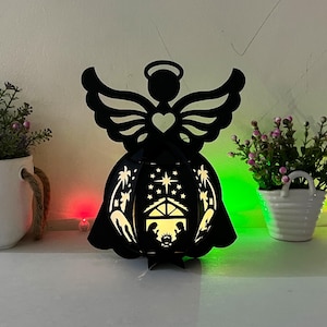 May include: Black silhouette of an angel holding a heart, with a nativity scene inside the angel's body. The scene is illuminated with a warm white light.