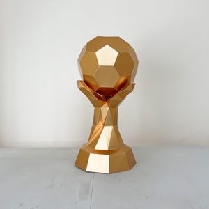Könnte beinhalten: Eine goldfarbene Trophäe mit geometrischem Design. Die Trophäe hat oben eine Fußballform, die von einer handähnlichen Struktur gehalten wird. Der Sockel hat eine vielseitige Form. Die Trophäe besteht aus glänzendem Metall.