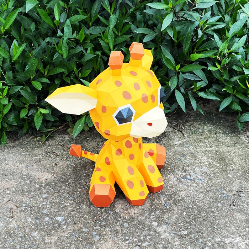May include: A yellow and orange papercraft giraffe with brown spots. The giraffe is sitting on a grey concrete surface.
