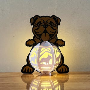May include: A white paper lantern with a silhouette of a French Bulldog on each side. The lantern is lit from the inside, casting a warm glow. A brown paper cutout of a French Bulldog is standing behind the lantern.