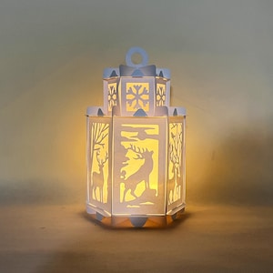 May include: A white paper lantern with a snowflake design on the top and a deer silhouette on the front. The lantern is lit from within, casting a warm glow.