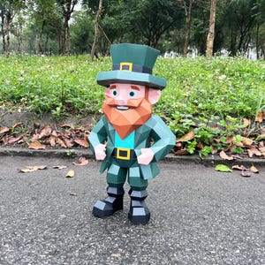 May include: A papercraft leprechaun figure in shades of green, with a red beard and a top hat. The figure has a geometric design and is standing on a paved surface. The background features greenery and trees.