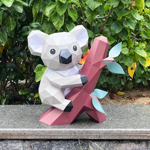 May include: A low-poly papercraft koala figure in shades of light gray and white with a dark gray nose and eyes. The koala is perched on a geometric, dark pink tree branch featuring several light blue paper leaves attached to the sides.