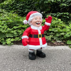 Può includere: Una figura di Babbo Natale in carta rossa e bianca con cintura e stivali neri. La figura è in piedi su una superficie grigia con fogliame verde sullo sfondo. La figura sta salutando con una mano.
