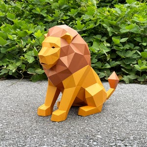 Può includere: Una scultura geometrica di leone di carta nei toni del giallo, arancione e marrone. Il leone è seduto con la coda arricciata, con sullo sfondo un fogliame verde.
