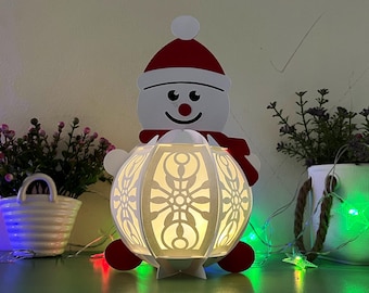 May include: A white snowman-shaped paper lantern with a red Santa hat and a white snowflake design. The lantern is lit from within, casting a warm glow.