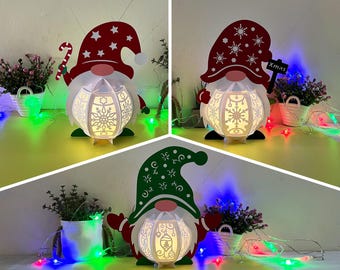 Pode incluir: Três luzes de Natal de papel em forma de gnomo com chapéus vermelho, verde e branco. Os gnomos estão segurando um bastão de caramelo, uma placa que diz "Xmas" e uma pequena árvore de Natal. Os gnomos são iluminados por luzes brancas e cercados por luzes de corda.
