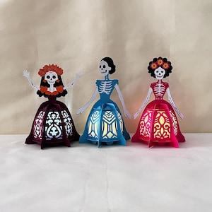 May include: Three decorative paper lanterns shaped like skeleton figures, each with a different dress colour: burgundy, blue, and pink. The lanterns have intricate cut-out designs and are illuminated from within, creating a warm glow.