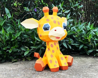 May include: A yellow papercraft giraffe with brown spots, sitting on a gray surface. The giraffe has large, blue eyes and a pink nose.
