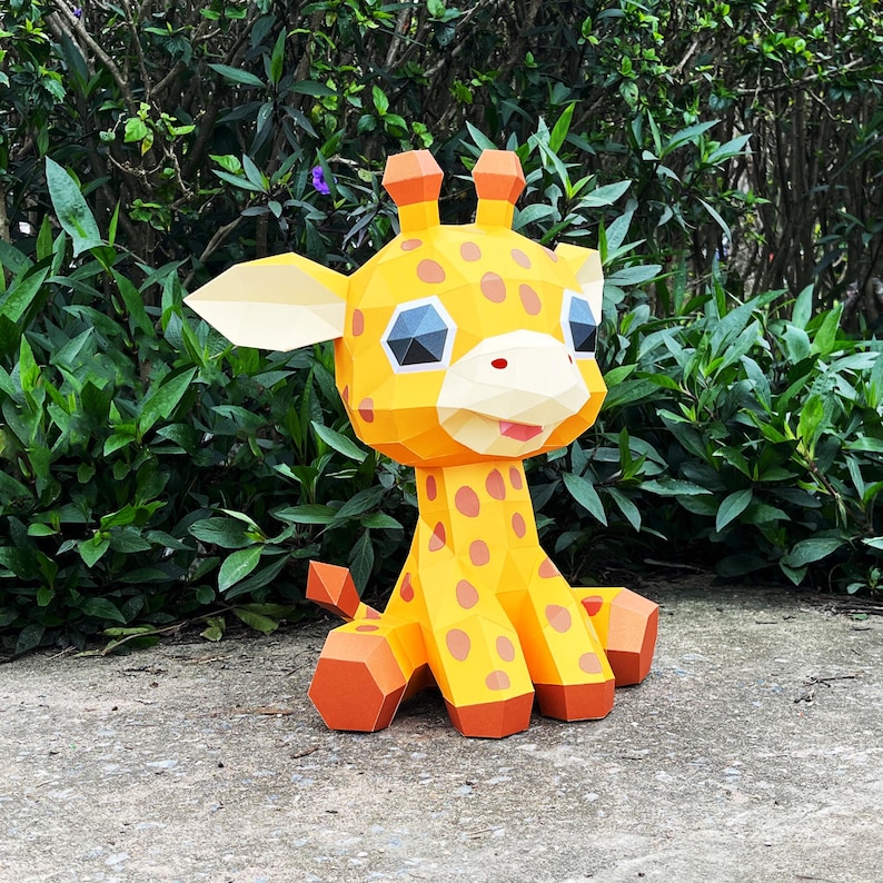 May include: A yellow papercraft giraffe with brown spots, sitting on a grey surface. The giraffe has large, blue eyes and a pink nose.