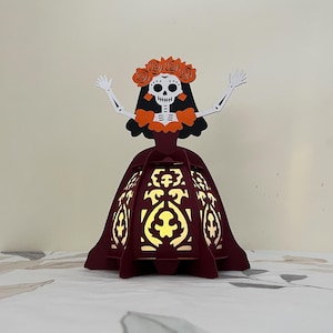 May include: A burgundy paper lantern in the shape of a Catrina, a female skeleton figure, with a floral crown and open arms. The lantern has intricate cut-out designs that allow light to shine through, creating a festive and decorative piece.