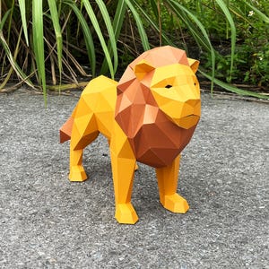 May include: A geometric paper lion sculpture in shades of yellow and brown stands on a gray surface. The lion has a detailed mane and body, with a polygonal design. The artwork is outdoors, with green foliage in the background.