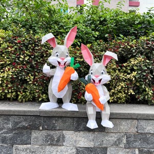 May include: Two papercraft rabbit figures in grey and white, each holding an orange carrot with green leaves. The bunnies have pink inner ears and are smiling. They are positioned in front of a green hedge.
