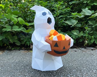 May include: A white geometric paper ghost figure holding a jack-o'-lantern candy bucket filled with candy corn. The ghost has black eye and mouth cutouts. The jack-o'-lantern is orange with a black face. The background is green foliage.