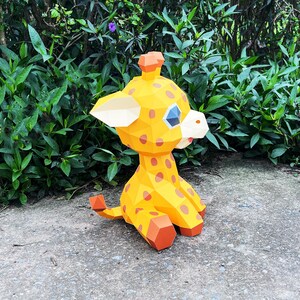 May include: A yellow and orange papercraft giraffe with brown spots. The giraffe is sitting on a grey concrete surface.
