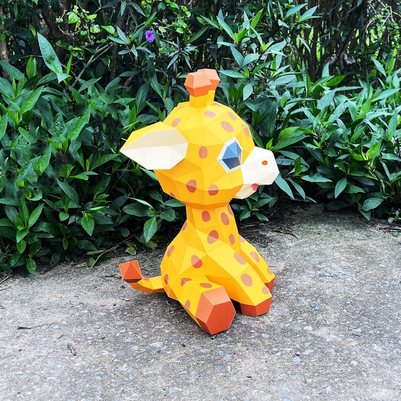 May include: A yellow and orange papercraft giraffe with brown spots. The giraffe is sitting on a grey concrete surface.