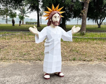May include: A papercraft sculpture of Jesus with outstretched arms. The figure is wearing a white robe, with a brown beard and hair, and a golden halo. The sandals are red and white. The sculpture is standing on a concrete surface with a grassy background.