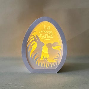 Puede incluir: Una lámpara de papel blanco en forma de huevo con un diseño 3D de dos conejos en su interior. La lámpara está iluminada desde el interior, proyectando un brillo cálido. El texto "Happy Easter" está escrito en la parte delantera de la lámpara.