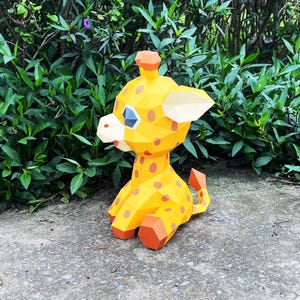 May include: A yellow and orange papercraft giraffe with black eyes and spots. The giraffe is sitting on a grey concrete surface.