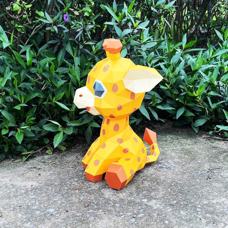 May include: A yellow and orange papercraft giraffe with black eyes and spots. The giraffe is sitting on a grey concrete surface.