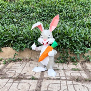 May include: A papercraft rabbit figure in shades of grey, holding a large orange and green carrot. The rabbit has pink inner ears and a smiling face. It stands on a tiled surface against a backdrop of green foliage.