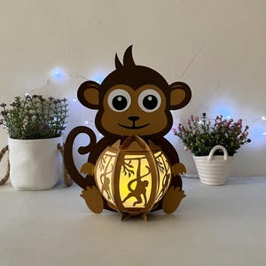 May include: A brown papercraft monkey holding a round, gold paper lantern with a light inside. The lantern has a cut-out design of monkeys swinging from vines.