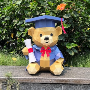 May include: A geometric papercraft teddy bear wearing a blue graduation cap and gown, holding a diploma with a red ribbon. The bear has a red bow tie and is set against a backdrop of green foliage and a stone surface.