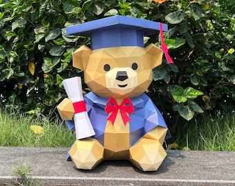 May include: A geometric papercraft teddy bear wearing a blue graduation cap and gown, holding a diploma with a red ribbon. The bear has a red bow tie and is set against a backdrop of green foliage and a stone surface.