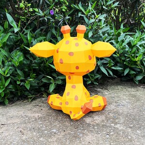 May include: A yellow papercraft giraffe with brown spots sitting on a grey concrete surface. The giraffe is facing away from the camera, with its head turned to the side.