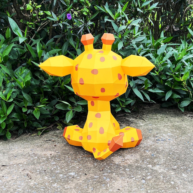May include: A yellow papercraft giraffe with brown spots sitting on a grey concrete surface. The giraffe is facing away from the camera, with its head turned to the side.