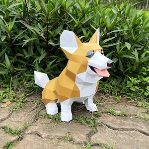 May include: A papercraft model of a corgi dog, made of white and brown paper. The dog is sitting on a stone path with green grass in the background.