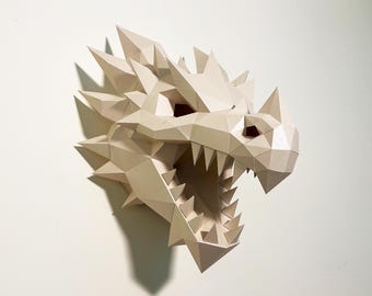 May include: A beige, geometric dragon head sculpture with sharp spikes and an open mouth revealing pointed teeth. The artwork is mounted on a white wall, casting a shadow. The dragon's eyes are visible within the head.