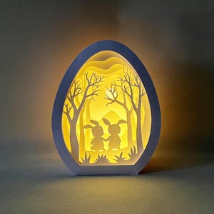 May include: A white papercraft egg-shaped lamp with a 3D scene of two bunnies in a forest. The lamp is lit from within, casting a warm glow.