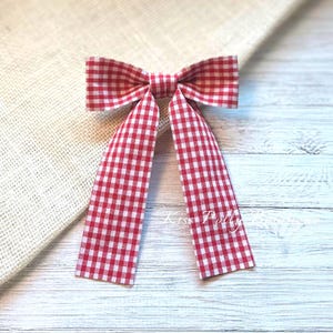 Puede incluir: Lazo de gingham rojo y blanco con un nudo central. El lazo está hecho de tela y tiene un patrón a cuadros.