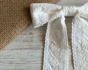 Lace Bloom: fermaglio per capelli in cotone bianco con fiocco ricamato a fiori e bordo smerlato.