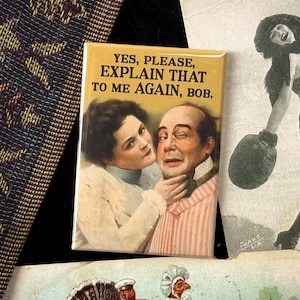 Op de afbeelding: Een rechthoekige magneet met de tekst "YES, PLEASE, EXPLAIN THAT TO ME AGAIN, BOB." toont een vintage illustratie van een vrouw en een man. De afbeelding is omgeven door vintage ephemera, waaronder een zwart-gouden textiel en een sepia-foto.