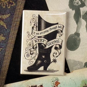May include: A rectangular magnet with a black and white illustration of a cowboy boot and the text "If you are going through hell, KEEP GOING." The magnet is surrounded by vintage ephemera, including a sepia-toned photograph of a woman.