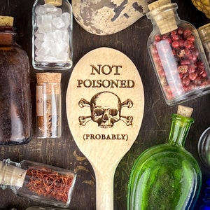 May include: Wooden spoon with a skull and crossbones design and the text "NOT POISONED (PROBABLY)". The spoon is surrounded by small glass bottles filled with various ingredients.