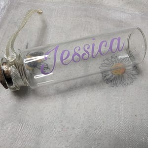 May include: A clear glass bottle with a cork stopper and a purple ribbon. The bottle has the name "Jessica" written on it in purple lettering. The bottle is sitting on a white, sparkly surface.