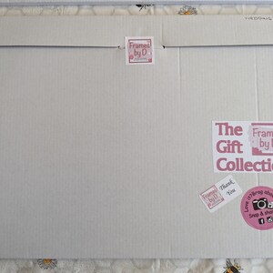 May include: A large, rectangular cardboard package with the text "WEDDING" printed on the top flap. The package features several stickers, including "Frames by D" and "The Gift Collection". A pink sticker encourages sharing on social media.